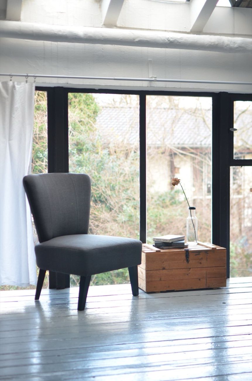 A grey armchair by a full-height window, painted wood floor, morning light